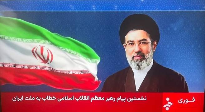 이란 국영TV가 12일(현지시간) 모즈타바 하메네이 최고지도자 명의의 첫 공식 성명을 방송하며 그의 사진과 이란 국기를 화면에 내보내고 있다. 성명은 영상이나 음성 메시지 없이 앵커가 대신 읽는 형식으로 전달됐다. 이란 국영TV 화면 캡처