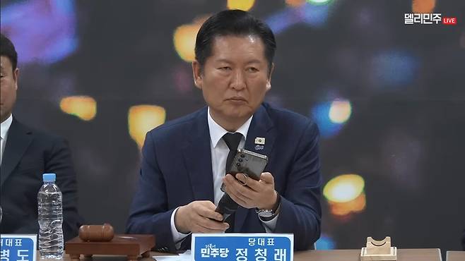 정청래 더불어민주당 대표가 23일 경남 김해시 봉하마을에서 주재한 현장 최고위원회의에서 노무현 전 대통령 논두렁 시계 관련 과거 보도 영상을 휴대전화로 재생하고 있다. 민주당 유튜브 채널 <델리민주> 영상 갈무리