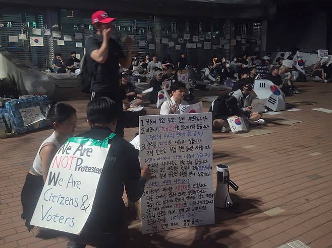 6일 오후 11시쯤 서울 송파구 잠실 투표소 앞. 집회 안내문 뒤로 야간 집회에 참여하는 시민들이 앉아 구호를 외치고 있다./김도연 기자